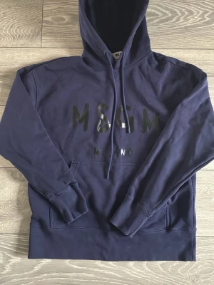 Mörkblå hoodie från MSGM Milano - Snygg mörkblå hoodie från MSGM Milano med stor huva och dragsko. Framsidan har ett svart MSGM-tryck och en klassisk känguruficka. Tillverkad i mjuk bomull, perfekt för en avslappnad streetwear-look. Använd endast ett få antal gånger, priset är ej hugget i sten, skriv vid minsta fundering. Nypris runt 2000.