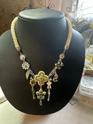 Pärlhalsband med blommor och berlocker - Unikt halsband med vita pärlor och flera detaljerade berlocker i form av blommor, löv och figurer i gult, blått och silver. Halsbandet har en bohemisk känsla med inslag av gnistrande stenar och olika former som ger en lekfull och kreativ look.