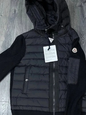 Svart pufferjacka från Moncler - Snygg svart pufferjacka från Moncler med stickade ärmar och vadderad kropp. Jackan har huva med dragsko, två fickor med tryckknappar och Moncler-logga på ärmen. Perfekt för kalla dagar och ger en sportig vibe. Passar både S och M