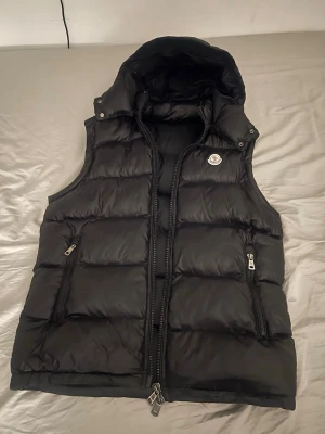 Svart dunväst från Moncler med huva - Snygg svart dunväst från Moncler med avtagbar huva och två dragkedjeförsedda fickor. Västen har klassisk Moncler-logga på bröstet och quiltad design. Insidan har etiketter och ett coolt serietidningsmönster. Perfekt för lager-på-lager och streetstyle.