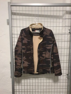Kamo vindjacka från Clique - Säljer en snygg vindjacka från Clique i storlek S med camouflage-mönster i brunt och grönt. Jackan har beige fleecefoder, hög krage och dragkedja framtill. Perfekt för dig som gillar streetwear och vill ha en unik look.