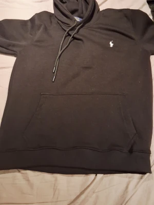 Svart hoodie från Polo Ralph Lauren - Klassisk svart hoodie från Polo Ralph Lauren med vit broderad logga på bröstet. Hoodien har huva med dragsko och en stor magficka framtill. Tillverkad i mjukt bomullsmaterial som känns skönt mot huden. Perfekt för en avslappnad och stilren look.
