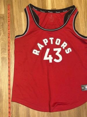 Röd Raptors basketlinne Siakam #43 - Snyggt rött basketlinne från Fanatics med Toronto Raptors tryck och nummer 43 på framsidan. På baksidan står det Siakam och samma nummer. Linnet har svarta och vita detaljer runt ärmhål och hals. Perfekt för basketfans som vill sticka ut.