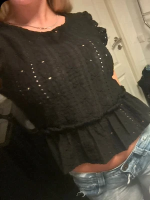 Svart broderad blus med volang - Säljer en svart blus med broderade detaljer och volangkant nedtill. Blusen har korta ärmar och ett lätt genombrutet mönster som ger en luftig känsla. Perfekt att styla med jeans eller kjol för en trendig look.