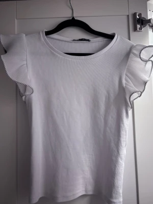 Vit ribbad topp med volangärmar - Slutsåld topp från zara som inte går att få tag i längre. Supersöt vit ribbad topp med volangärmar och kontrastsömmar i svart längs volangkanten. Perfekt för dig som gillar en feminin och trendig stil. Rund halsringning och figurnära passform.