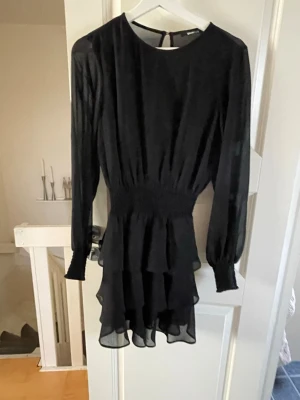 Svart volangklänning med lång ärm - Svart klänning med långärmad design och volangdetaljer på kjolen. Klänningen har en markerad midja och är tillverkad i ett lätt, transparent material som ger ett snyggt fall. Perfekt för dig som gillar stilrena och trendiga plagg.