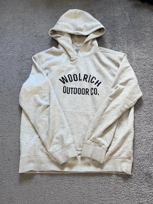 Grå hoodie från Woolrich Outdoor Co. - Ljusgrå hoodie från Woolrich Outdoor Co. med svart tryck på bröstet. Klassisk modell med huva och ribbade muddar vid ärmslut och nederkant. Tillverkad i mjukt sweatshirtmaterial som känns skönt mot huden. Perfekt för en avslappnad och sportig stil.