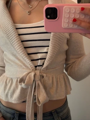 Beige kofta med knytband från H&M - Supersöt beige kofta från H&M med bred ribbad krage, knytband i midjan och volangkant nedtill. Långärmad och i mjukt stickat material som känns riktigt skönt mot huden. Perfekt att slänga över en topp för en chill och snygg look.