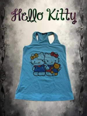 Blå Hello Kitty linnetopp - Söt blå linnetopp från Hello Kitty med tryck av två Hello Kitty-figurer och en nalle på framsidan. Toppen är ärmlös med brottarrygg och har en slim passform. Perfekt för dig som älskar gulliga motiv och vill ha en bekväm topp till sommaren.