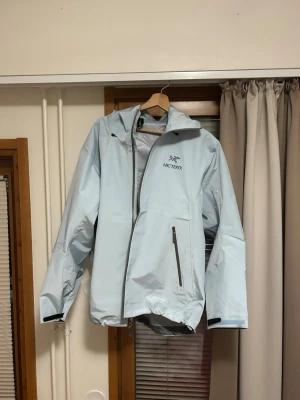 Ljusblå regnjacka från Arc'teryx - Snygg ljusblå regnjacka från Arc'teryx med huva och dragkedja framtill. Jackan har en bröstficka med vattentät dragkedja och justerbara muddar. Tillverkad i slitstarkt, vattenavvisande material som håller dig torr under regniga dagar.