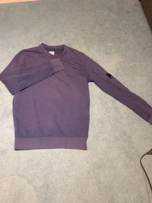 Marinblå sweatshirt från C.P. Company - Säljer en sweatshirt från C.P. Company med rund hals och ribbade muddar. Tröjan har en ikonisk detalj med glaslins på ena ärmen och är tillverkad i mjuk bomull. Perfekt för en avslappnad och stilren look.