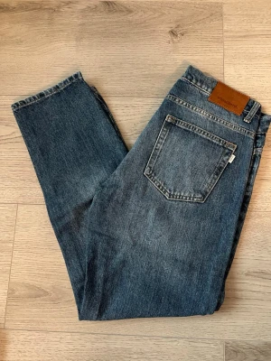Vintage blå raka jeans från Woodbird - Blå raka jeans från Woodbird med rak passform och fem fickor. Jeansen har en vintage tvätt och brunt läderpatch bak i midjan. Tillverkade i slitstark denim med knappgylf och tydliga sömmar. Perfekta för en avslappnad och tidlös stil.