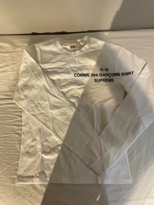 Vit långärmad topp Supreme x Comme des Garçons - Vit långärmad topp från Supreme x Comme des Garçons med svart tryck på bröstet. Klassisk rund halsringning och rak passform. Tillverkad i mjuk bomull som känns skön mot huden. Perfekt för dig som gillar streetwear och minimalistisk stil.