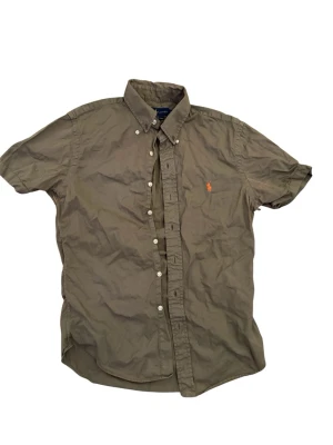 Olivgrön kortärmad skjorta Ralph Lauren - Kortärmad skjorta i olivgrönt från Ralph Lauren med klassisk button-down krage och orange broderad logga på bröstet. Slim fit-modell i lätt twill-bomull som känns tunn och luftig. Perfekt för dig som gillar stilrena och tidlösa plagg. Hör av er vid fler frågor eller funderingar, pris kan diskuteras.