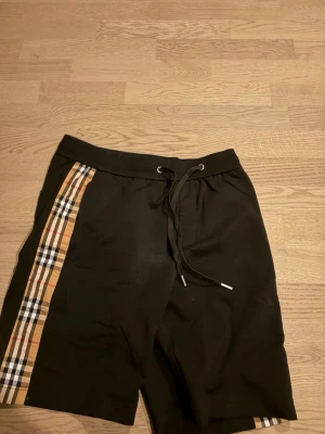 Svarta Burberry shorts  - Svarta shorts från Burberry med klassiska rutiga paneler längs sidorna och snörning i midjan. Bakficka med Burberry-logga. Perfekta för en trendig och avslappnad stil. Det är äkta shorts!