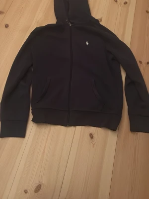 Mörk blå hoodie från Polo Ralph Lauren - Mörk blå hoodie med dragkedja från Polo Ralph Lauren. Klassisk design med två fickor framtill, liten broderad logga på bröstet och huva. Perfekt för en avslappnad och stilren look. Passar dig som är runt 170 cm