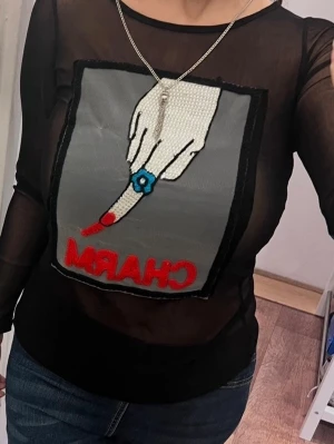 Vår Svart mesh topp - Säljer en svart, transparent mesh topp med lång ärm och ett stort grafiskt tryck framtill som föreställer en hand med blå ring och texten 'CHARM' i rött. Toppen har en tight passform och är perfekt för dig som vill sticka ut.