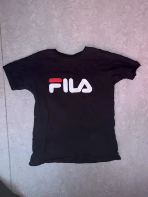 Svart FILA t-shirt med logga - Säljer en svart t-shirt från FILA med stor vit och röd logga på bröstet. Klassisk rund hals och korta ärmar. Perfekt för dig som gillar sportig och enkel stil.