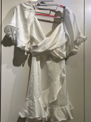 Vit klänning  med volanger - Säljer en superfin vit kortklänning med volangdetaljer.💞💞 Klänningen har vida, korta ärmar och ett lätt, flödande tyg som ger en romantisk känsla. Den här knyt så att man kan justera baksidan, göra rosett. Perfekt för sommarens alla tillfällen och studenten!!! 💞