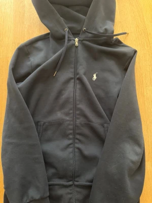 Svart hoodie från Polo Ralph Lauren - Klassisk svart hoodie från Polo Ralph Lauren med dragkedja framtill, fickor och justerbar huva. Ikoniska loggan broderad på bröstet. Tillverkad i mjuk bomullsblandning som känns skön mot huden. Perfekt för en avslappnad och stilren look.