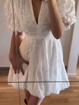 Vit kort klänning från Zara - Supersöt vit kort klänning från Zara med puffärmar, djup v-ringning och broderade blommor. Klänningen har en markerad midja och ballongformad kjol som ger en trendig siluett. Perfekt för sommarens alla tillfällen o studneten!