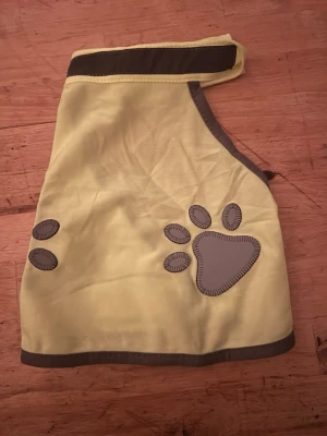 Gul reflexväst för hund - Säljer en gul reflexväst för hund med grå kantband och reflexdetaljer i form av tassavtryck. Västen har kardborreband vid hals och mage för enkel på- och avtagning. Perfekt för promenader i mörkret så din hund syns bättre.