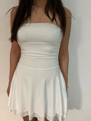 Vit axelbandslös kort klänning - Säljer en vit, axelbandslös kort klänning med figurnära överdel och utställd kjol. Klänningen har en enkel och stilren design som ger en fräsch look. Perfekt för varma dagar och sommarens alla tillfällen. Använd en gång