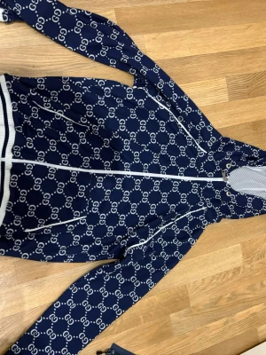 Mörkblått set med GG-mönster från Gucci - Snyggt tvådelat set från Gucci i mörkblått med vitt GG-monogram över hela plagget. Setet består av långärmad zip-jacka och matchande byxor med vita detaljer. Materialet ser ut att vara mjukt och stretchigt, perfekt för en trendig look.