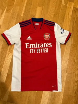 Arsenal matchtröja röd/vit Adidas - Säljer en Arsenal matchtröja från Adidas i rött och vitt med klubbmärke och sponsortryck på bröstet. Tröjan har blå detaljer på axlarna, korta ärmar och är tillverkad i ett lätt, ventilerande material. Perfekt för fotbollsträning eller att bära till match.