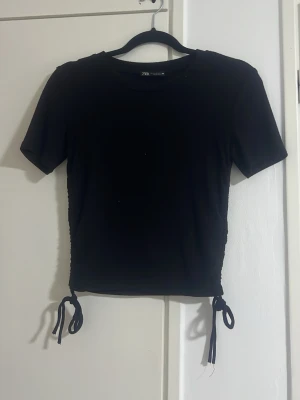 Svart topp med snörning från Zara - Snygg svart topp från Zara med korta ärmar och trendig snörning i sidorna som kan justeras.