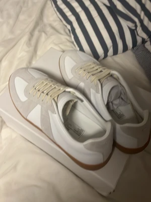 Vita sneakers från Maison Margiela - Säljer ett par stilrena vita sneakers från Maison Margiela med beigea mockadetaljer och klassisk gummisula. Skorna har vita snören och en retroinspirerad design som passar perfekt till streetwear eller casual outfits.