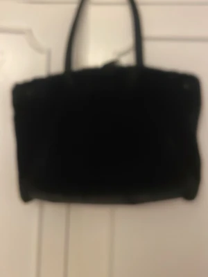 Svart fluffig handväska - Säljer en svart handväska med fluffig, pälsliknande yta och två handtag. Väskan har en rektangulär form och känns riktigt trendig och mysig. Perfekt accessoar för dig som vill sticka ut med en unik och cool look.