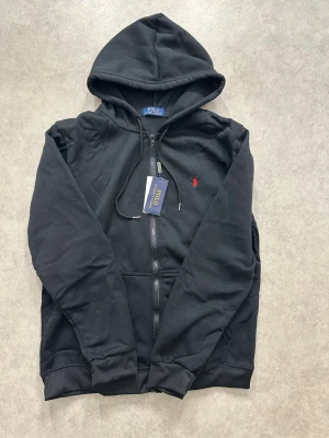 Svart hoodie från Polo Ralph Lauren - Klassisk svart hoodie med dragkedja från Polo Ralph Lauren. Hoodien har justerbar huva, fickor framtill och den ikoniska röda logotypen broderad på bröstet. Tillverkad i mjukt bomullsmaterial för en skön och avslappnad look.