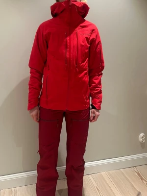 Röd skidjacka från Norrøna - Röd skidjacka från Norrøna med huva och flera dragkedjeförsedda fickor. Jackan har en sportig passform, justerbar huva och detaljer i svart på insidan. Perfekt för utomhusaktiviteter och skyddar mot vind.