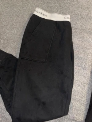 Svarta mjukisbyxor Calvin Klein - Svarta mjukisbyxor från Calvin Klein med bred, vit resår i midjan där loggan syns tydligt. Byxorna har raka ben och djupa fickor på sidorna. Tillverkade i mjukt och bekvämt material, perfekta för chill dagar eller när du vill ha det extra skönt.