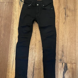 Svarta skinny jeans från Nudie Jeans - Svarta jeans från Nudie Jeans med klassisk femficksdesign och smal passform. Byxorna har svarta bakfickor med Nudie Jeans broderi och logga, samt silverfärgade knappar. Perfekta för dig som gillar en stilren och enkel look.