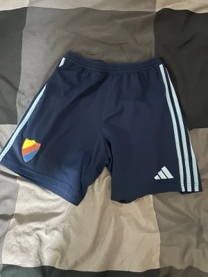 Blå Adidas Djurgården shorts - Mörkblå sportshorts från Adidas med ljusblå ränder längs sidorna och Djurgårdens IF klubbmärke på ena benet. Tillverkade i ett lätt och snabbtorkande material, perfekta för träning eller match. Elastisk midja för skön passform.