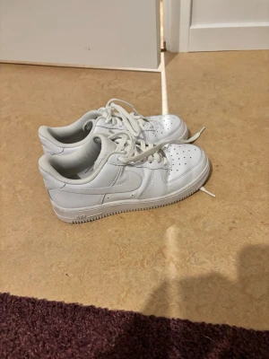 Nike Air Force 1 vita sneakers - Klassiska vita Nike Air Force 1 sneakers i lågt utförande. Skorna har en stilren design med perforerad tå, vita skosnören och Nike-logga på sidan och hälen. Tillverkade i läder med en robust sula som ger skön komfort och streetkänsla.