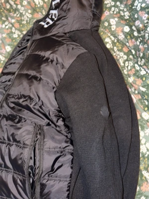 Svart pufferjacka från Moncler - Snygg svart pufferjacka från Moncler med quiltad design och stickade ärmar. Jackan har huva med Moncler-logga, dragkedja framtill och sidofickor. Materialet är en mix av glansigt nylon och stickad textil på ärmarna. Perfekt för kalla vinterdagar.