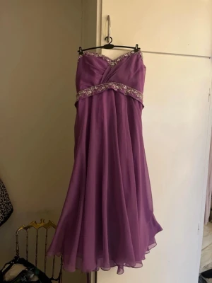 Lila långklänning med spetsdetaljer - Lila långklänning med v-ringad överdel och glittriga spetsdetaljer vid byst och midja. Klänningen har omlottkänsla framtill och snörning i ryggen för justerbar passform. Flödande kjol i tunt, svepande material som ger en elegant look.