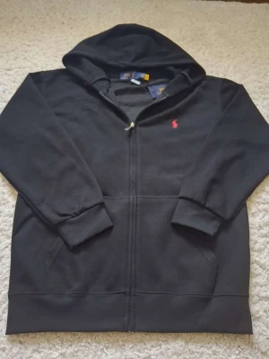 Svart hoodie från Polo Ralph Lauren - Klassisk svart hoodie från Polo Ralph Lauren med dragkedja framtill och huva. Ikoniska röda loggan broderad på bröstet. Tillverkad i mjukt bomullsmaterial och har ribbade muddar vid ärmar och nederkant. Perfekt för en avslappnad och stilren look.