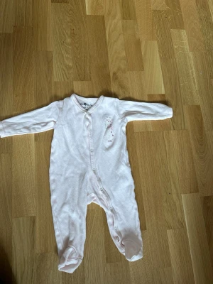 Ljusrosa pyjamas med giraffmotiv - Supersöt ljusrosa pyjamas från So Cute i storlek 68 cm (3-6 månader). Helkroppsmodell med långa ärmar och fötter, tryckknappar framtill och ett gulligt giraffmotiv på bröstet. Perfekt för mysiga nätter och lugna stunder hemma.