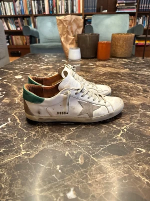 Golden Goose sneakers - Säljer ett par vita Golden Goose sneakers med grå stjärna på sidan och grönt parti vid hälen. Skorna har klassisk låg profil, vita snören och slitna detaljer för en cool look. Perfekta för dig som gillar streetstyle och trendiga märken.