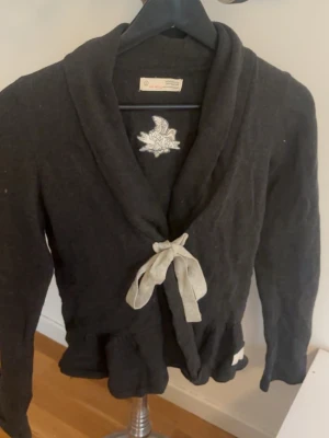 Svart kofta med knytband Odd Molly - Snygg svart kofta från Odd Molly med grått knytband i midjan och broderad logga på insidan. Koftan har lång ärm, sjalkrage och volangkant nedtill. Perfekt för lager på lager och en avslappnad men stilren look.