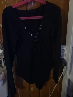 Svart långärmad body med snörning - Snygg svart body med långa ärmar och djup V-ringning framtill. Framsidan har cool snörningsdetalj med metallöljetter som ger en trendig look. Perfekt att styla med jeans eller kjol för en edgy outfit.