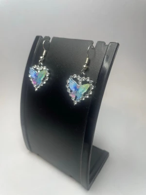 Hjärtfomade örhängen med fjäril - Säljer ett par örhängen i form av hjärtan med en glittrig fjäril i mitten. Örhängena har en silverfärgad kant dekorerad med små pärlliknande detaljer och fjärilen skimrar i blått, grönt och lila. Perfekt accessoar för dig som gillar unika smycken.