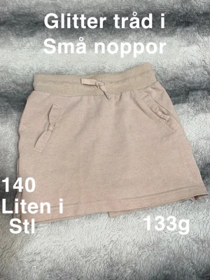 Rosa kjol med glittertråd - Söt rosa kjol med diskret glittertråd och ribbad midja med knytband. Kjolen har två fickor framtill med små rosetter som detalj. Perfekt för en trendig och avslappnad look.🌸 Små noppor. Glittertråd. Står 140 men upplever som liten i storlek.