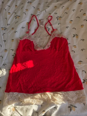 Rött linne med spetsdetaljer - S - Säljer ett skrikrosa linne med tunna axelband och vacker vit spets vid urringning och nederkant. Linnet har en feminin passform och spetsdetaljer även på ryggen. Perfekt för dig som vill ha något både bekvämt och snyggt till sommaren.