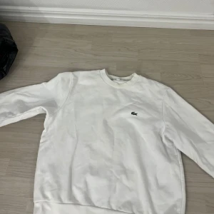 Vit sweatshirt från Lacoste - Klassisk vit sweatshirt från Lacoste med rund halsringning och det ikoniska gröna krokodilmärket broderat på bröstet. Tröjan har ribbade muddar vid ärmslut och nederkant. Perfekt för en clean och stilren look.