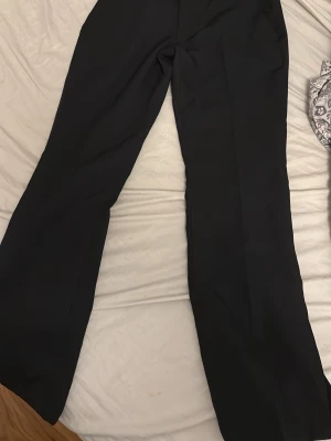 Svarta kostymbyxor med raka ben - Säljer ett par klassiska svarta kostymbyxor med raka ben och hög midja. Byxorna har bälteshällor och sidfickor, perfekta för en clean och stilren look. Materialet är slätt och ger en dressad känsla.